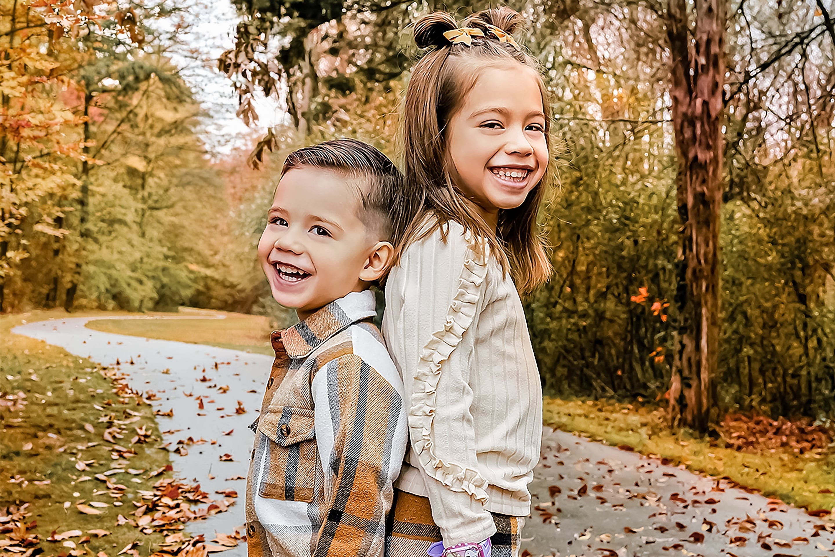 Brother and sister laughing back-to-back on a leaf-covered path in fall